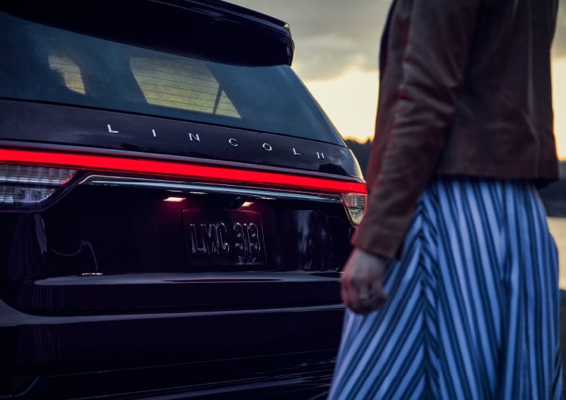 A person is shown near the rear of a 2024 Lincoln Aviator® SUV as the Lincoln Embrace illuminates the rear lights | Lincoln Demo 1 in Wooster OH