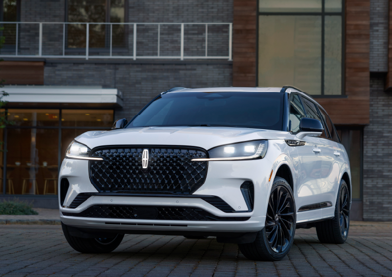 A white 2025 Lincoln Aviator® SUV is shown parked outside a modern building with gray brick and wood accents. | Lincoln Demo 1 in Wooster OH